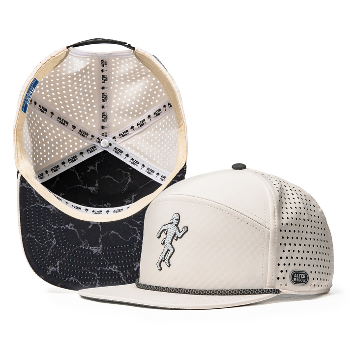 Alter Ego Drifter Splash Ivory Mummy running hat with an ivory design, flat bill, and running mummy graphic shown alongside an interior view featuring a black storm-patterned underbill and breathable perforated lining.