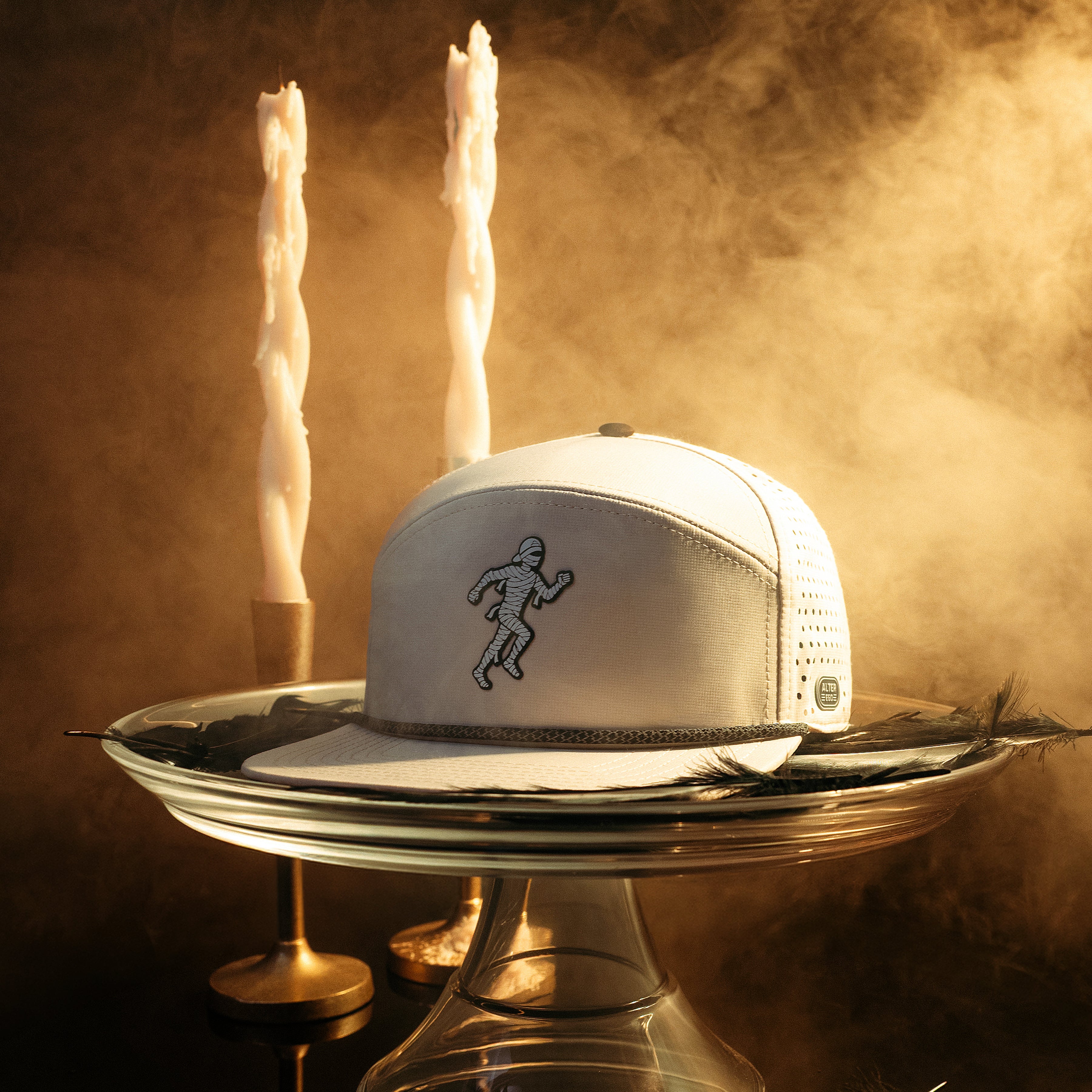 Alter Ego Drifter Splash Ivory Mummy running hat with an ivory design and running mummy graphic displayed on a glass pedestal in a Halloween-inspired scene with candles, smoke, and dark feathers.