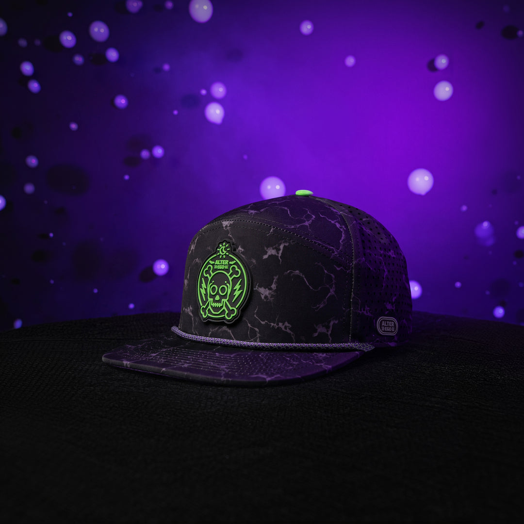 Drifter Splash Toxic Storm performance hat with a black lightning pattern, flat bill, neon green skull graphic, and breathable laser-cut side panels against a glowing purple background.